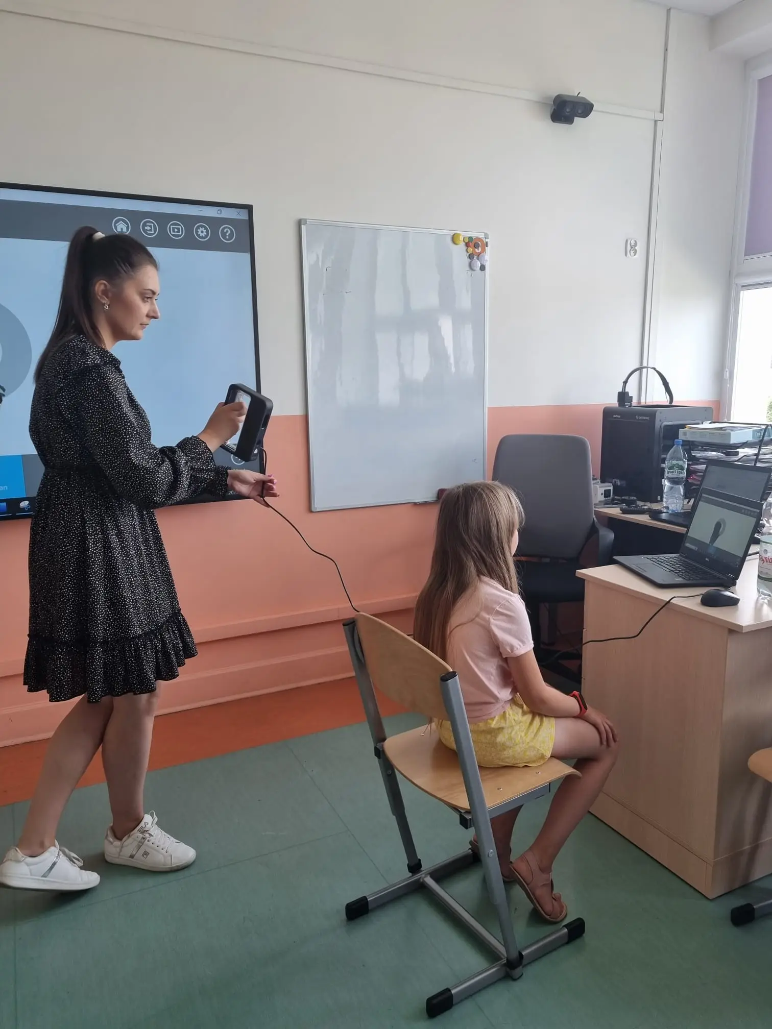 Prowadząca skanuje ręcznym skanerem 3D siedzącą dziewczynkę